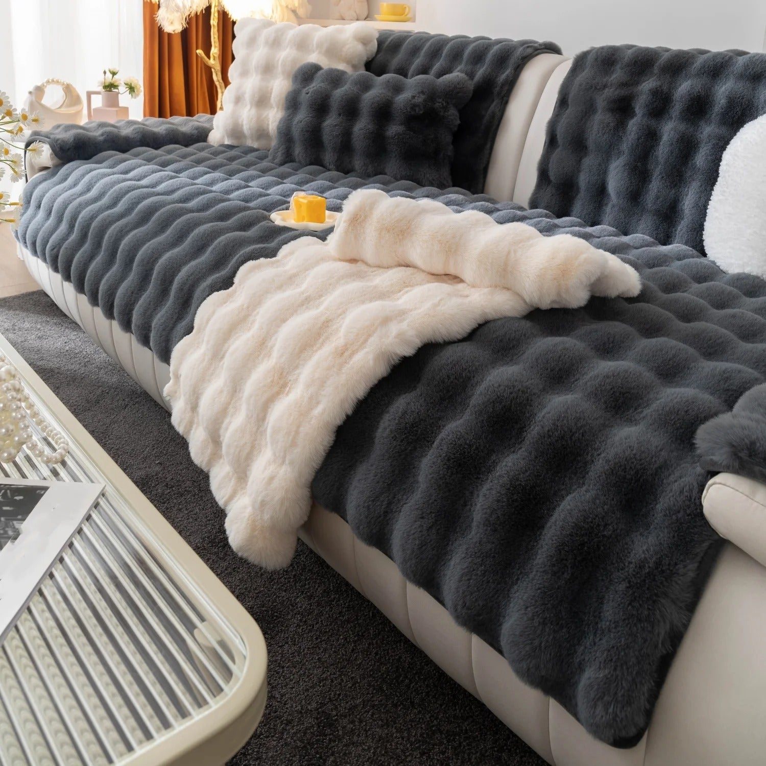 Een luxe woonkamer met een grote, zachte grijze bank bedekt met lagen zachte, getextureerde Act Design Shop Wagner - Fluffy Bankhoes Universeel dekens in donkergrijs en crème tinten. Een strakke, moderne